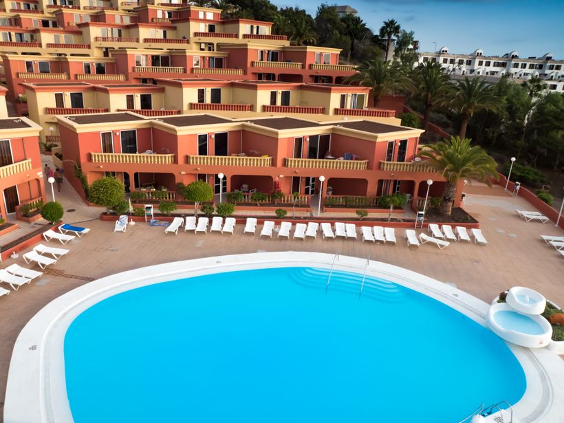 Aerial view of terracotta-coloured resort apartments with palm trees around a curved blue swimming pool and sun loungers