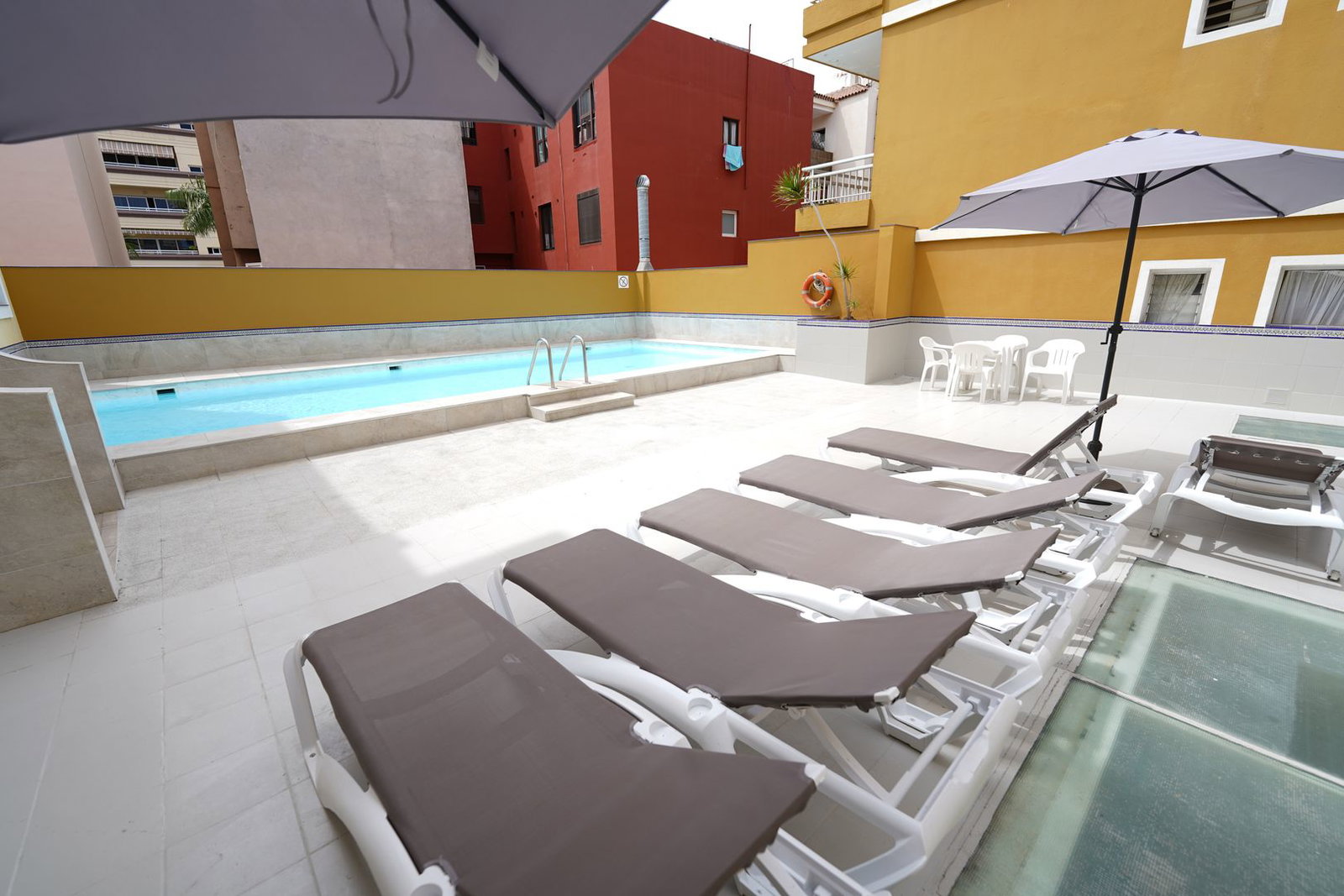 Outdoor hotel pool terrace with row of brown sunloungers and parasols, white plastic table and chairs, lifebuoy and surrounding apartment buildings