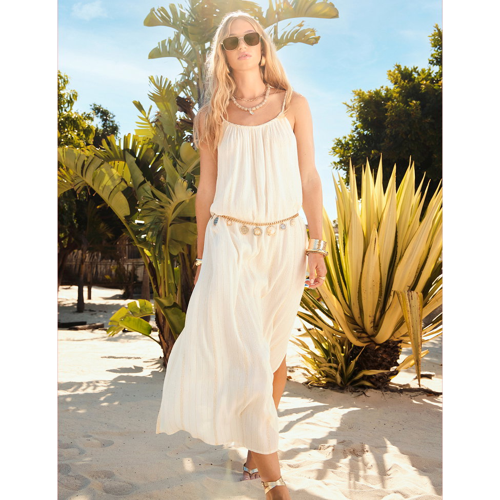 Model wearing a flowing cream maxi dress walks across a sunlit tropical beach surrounded by palm trees and exotic plants. Styled with sunglasses, layered jewellery and metallic sandals, the look creates an effortless holiday-ready outfit.