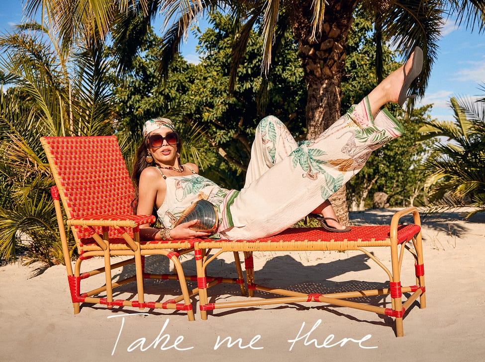 Woman relaxing on a red woven sun lounger beneath palm trees, wearing a tropical print co-ord set and sunglasses on a sunny beach. White script text reads "Take me there."