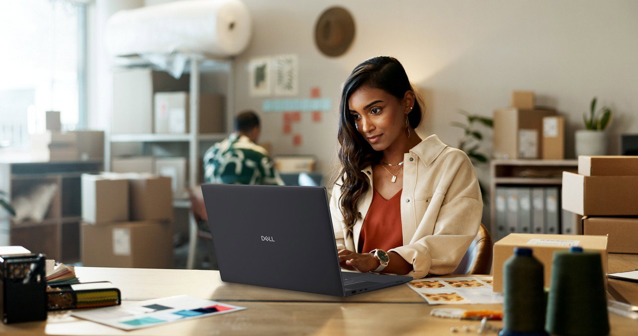 Woman working on a Dell laptop at a creative studio workspace surrounded by boxes, fabrics and design materials.