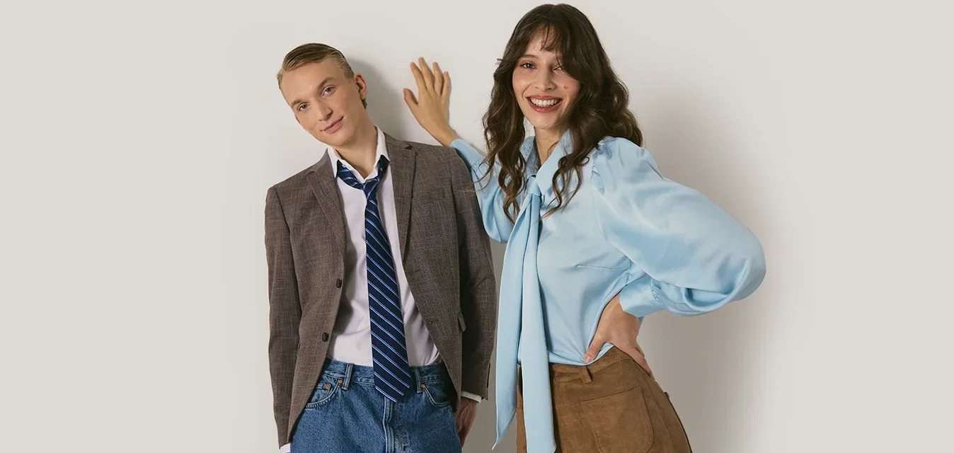 Twee modellen staan tegen een lichte achtergrond. De één draagt een grijze blazer, een blauwe stropdas, een wit shirt en spijkerbroekshorts. De andere heeft een lichtblauwe blouse en een bruine rok aan.