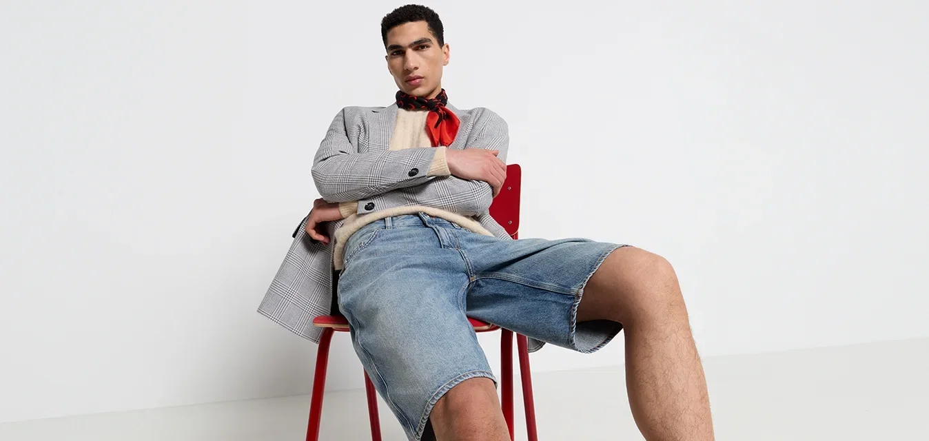 Grijze geruite blazer over een beige sweater, denim shorts en een rode sjaal. Zittend op een rode stoel tegen een effen witte achtergrond.