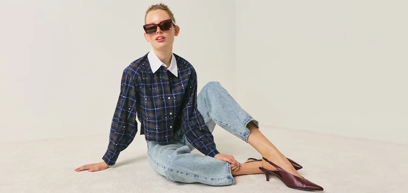 Frau mit Sonnenbrille, einem dunklen karierten Hemd, hellblauen Jeans und violetten Absätzen, sitzt auf dem Boden vor einem schlichten neutralen Hintergrund.
