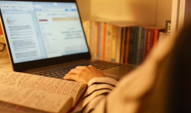 Person studying at a desk with handwritten notes in a notebook and a laptop displaying text, with a shelf of books in the background.