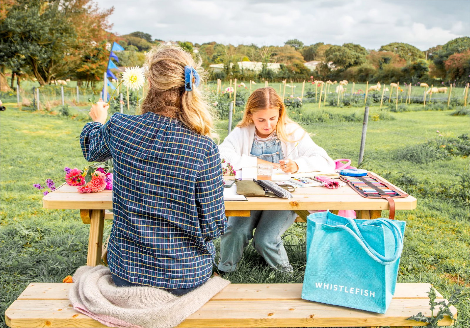 Members of the Whistlefish creative team sketching and painting outdoors at a picnic table surrounded by flowers and countryside views.