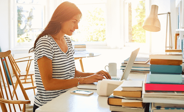 Woman sitting at desk typing on laptop surrounded by books.