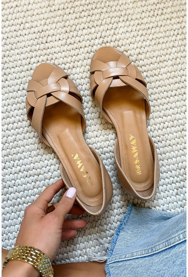 Pair of tan woven leather flat sandals on a cream textured rug, hand with pale pink manicure holding one beside denim hem