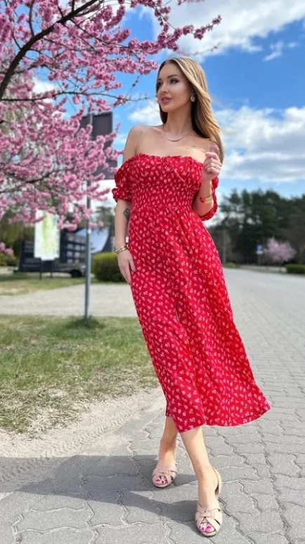 Woman in red off-shoulder floral dress walking past pink cherry blossoms on a sunny spring day with blue sky