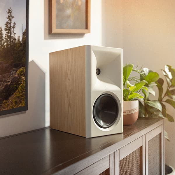 Klipsch Seven II powered speaker, in Red Oak finish, sitting on a dark wooden tv caninet with potted plants around it. 