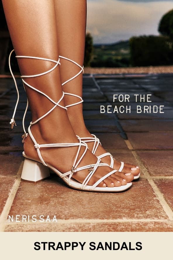 A close-up photo shows a person's foot wearing white "NERISSA" strappy block heel sandals featuring delicate coastal charms. Overlaid white text on the right reads "FOR THE BEACH BRIDE", and the bottom text reads "STRAPPY SANDALS".