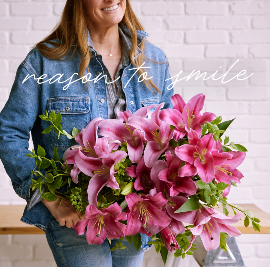 Farmgirl Flowers campaign image showing a smiling person in a denim jacket holding a large pink lily bouquet with greenery. White script text reads “reason to smile,” with the Farmgirl Flowers logo near the top.