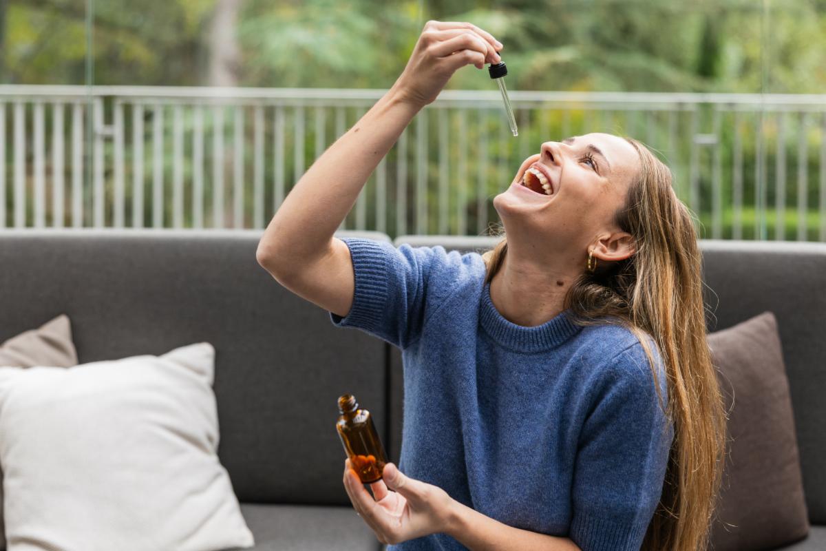 Young woman laughing while taking cbd oil with a dropper sitting on a comfortable sofa on her home's terrace