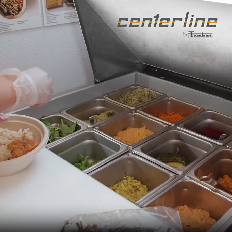 Centerline by Traulsen refrigerated prep unit with stainless steel food pans holding fresh toppings as a gloved worker assembles a bowl.