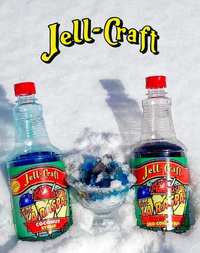 Jell‑Craft sno‑cone syrups in coconut and grape flavors displayed in snow alongside a cup of colorful shaved ice, with bold Jell‑Craft branding above.