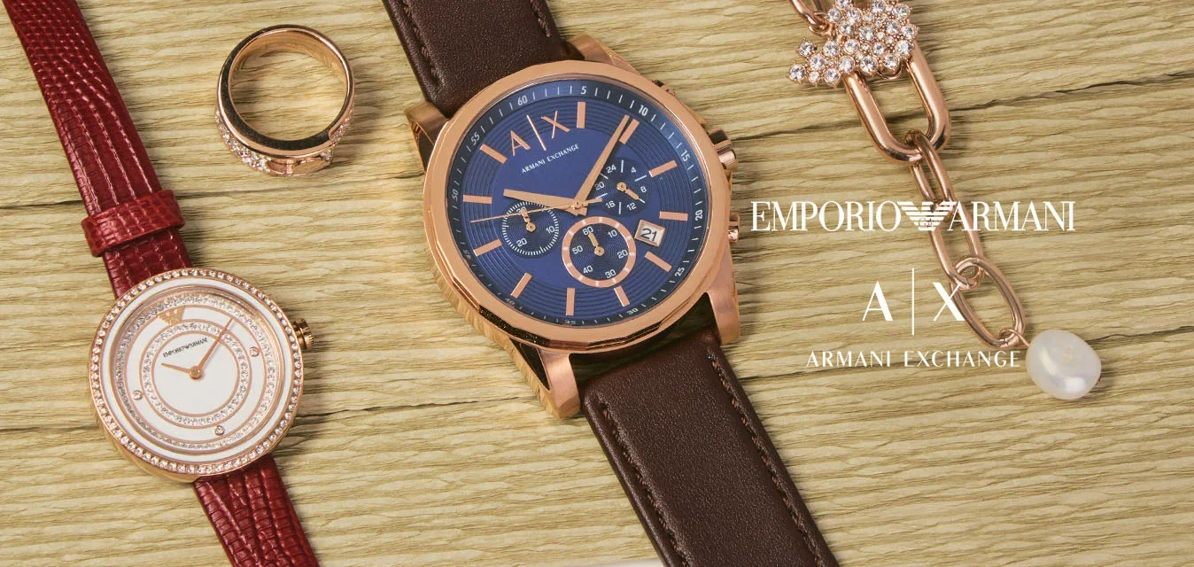 Zwei Armbanduhren, ein Goldring und eine Goldkette mit einem Schmetterlingsanhänger und einer Perle sind auf einer beigen, strukturierten Fläche angeordnet.