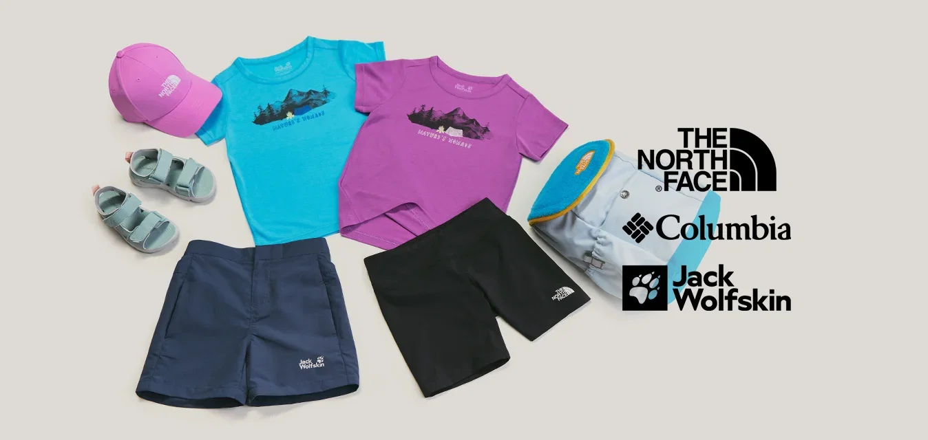 Zwei T-Shirts, zwei Shorts, Sandalen, eine pinke Kappe und ein blauer Rucksack auf hellem Hintergrund angeordnet.