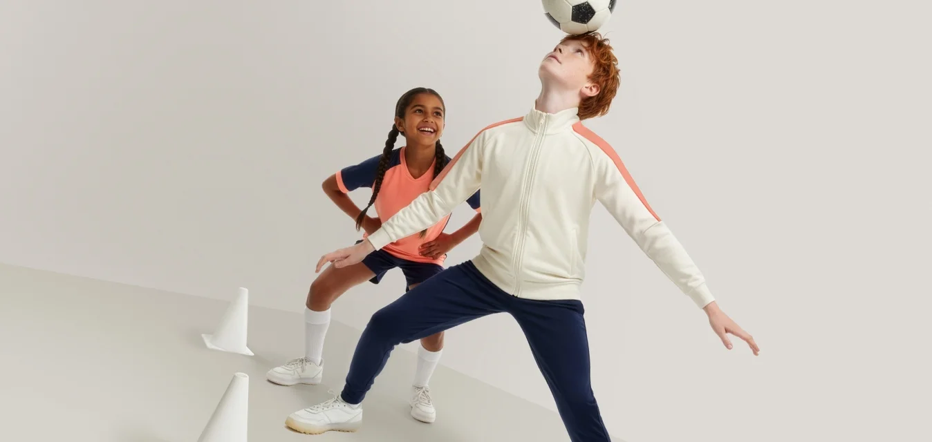 Ein Junge balanciert einen Fußball auf dem Kopf, während ein Mädchen zuschaut und lächelt; beide tragen Sportbekleidung und stehen in der Nähe von Trainingshütchen.