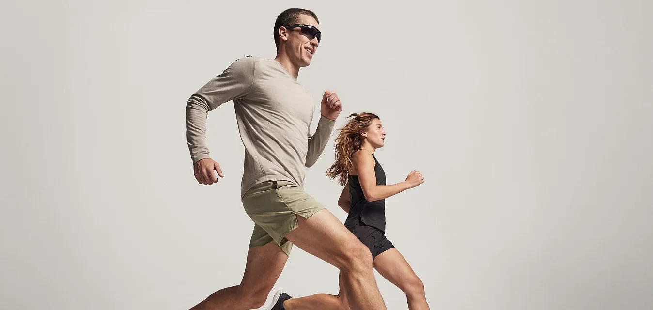 Twee volwassenen, een man in een korte broek en zonnebril en een vrouw in een zwart sportoutfit, rennen naast elkaar tegen een effen achtergrond.