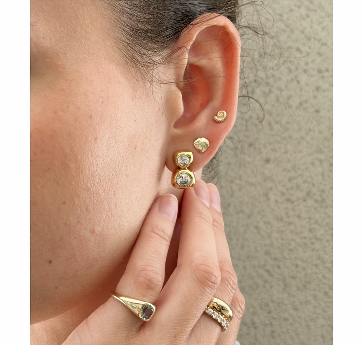 Close-up of a woman's ear styled with Mejuri gold drop studs, shell and ammonite studs, with stacked gold rings on her hand.