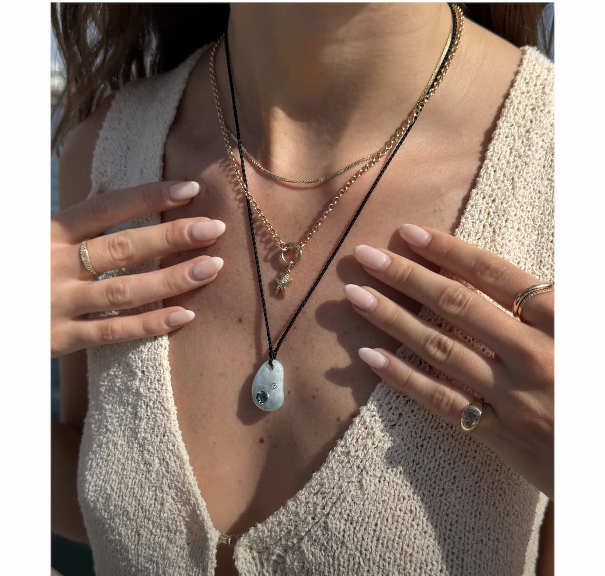 Woman's neckline layered with Mejuri gold chain necklaces, a stone pendant on a dark cord, and stacked rings.