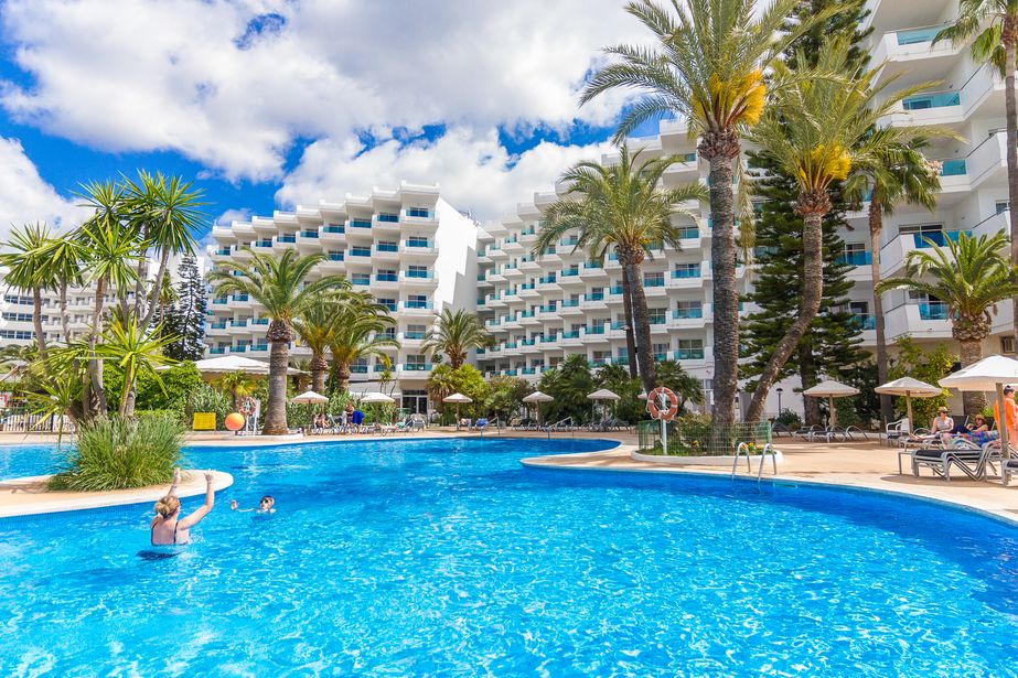 Sunlit resort hotel with large blue swimming pool, palm trees, sunloungers and guests swimming under balconies