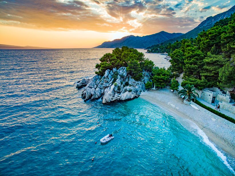 Sunset over turquoise Adriatic sea with pebble beach, rocky pine-topped islet and small inflatable boat near shore