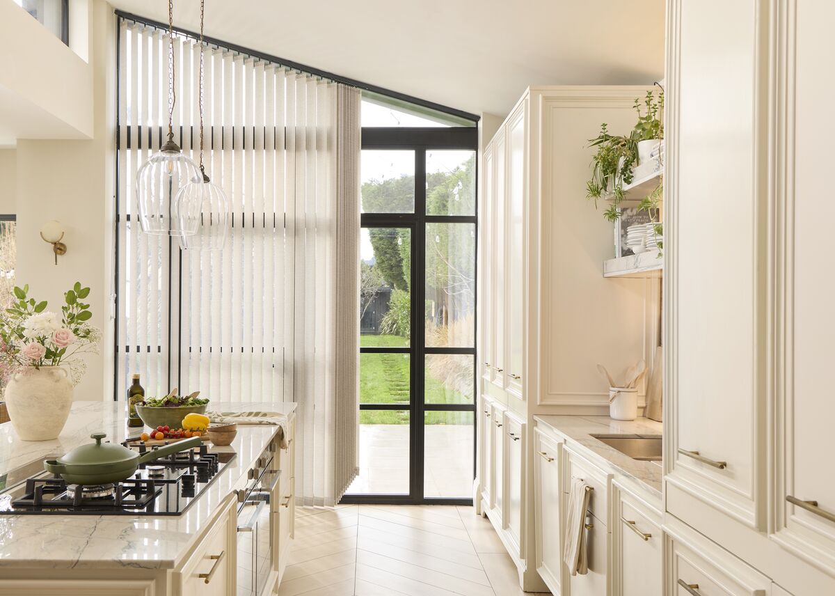 Dining room with wide window dressed in soft blue Vertical blinds, filtering daylight over a wooden table set with ceramics and greenery.