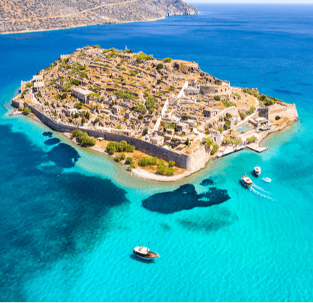 Aerial view of a rocky island with ancient ruins surrounded by turquoise waters and boats.