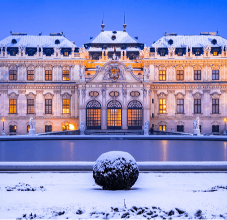 A grand historic building illuminated at dusk, surrounded by snow and a frozen pond.