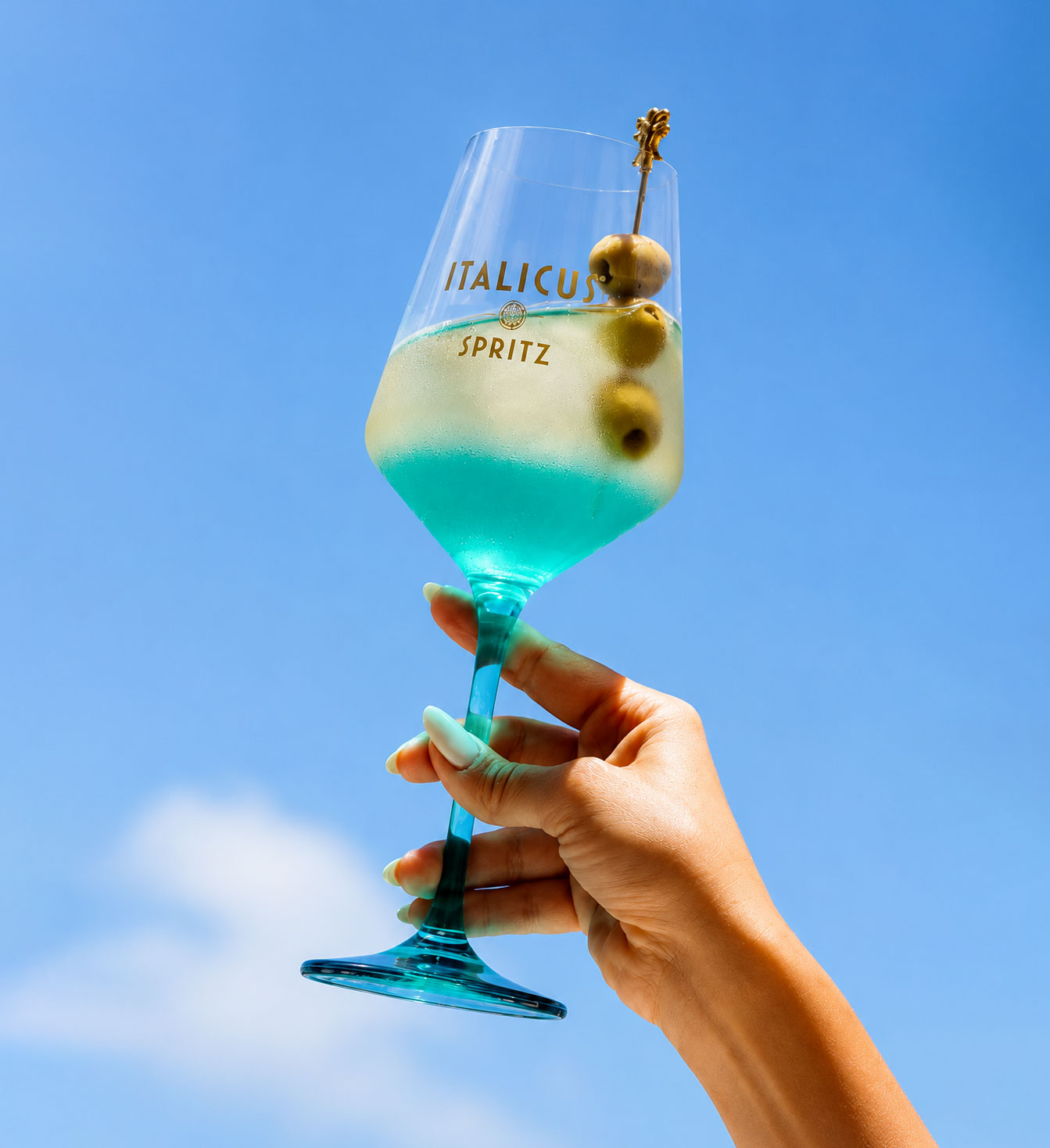 Hand holding turquoise-stemmed glass of Italicus Spritz with blue-green cocktail and three olives on a skewer against a clear blue sky