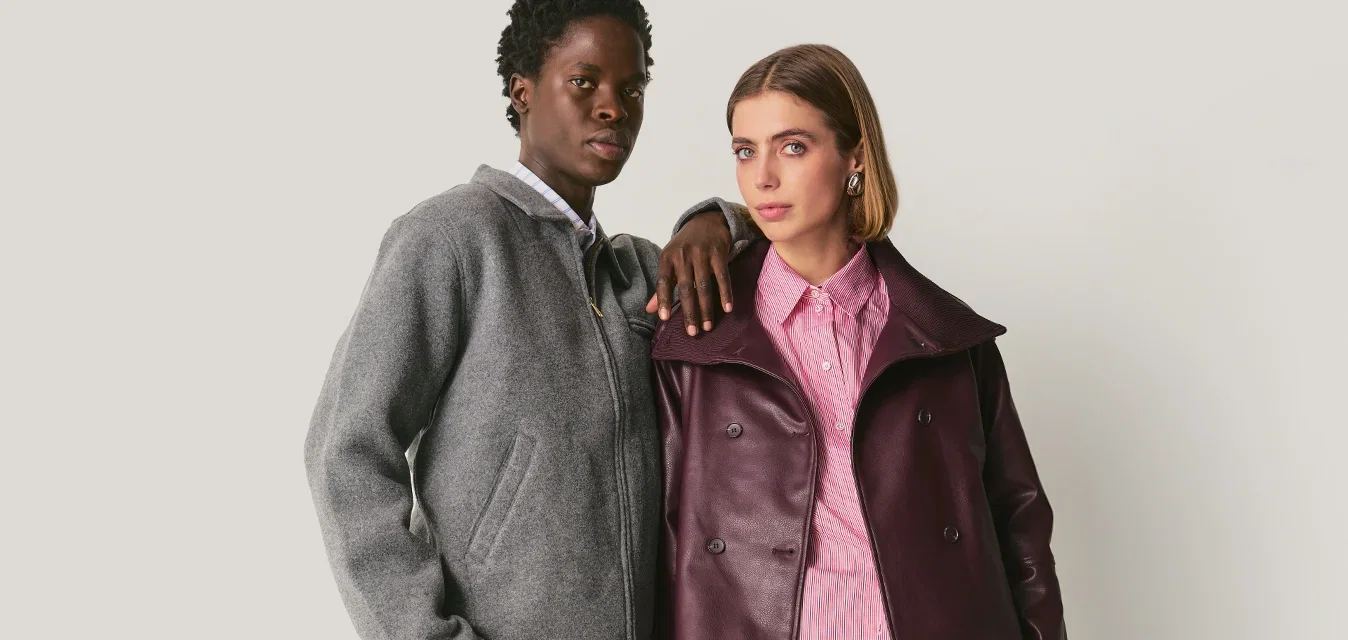 Twee mensen staan dichtbij elkaar; de één draagt een grijze jas en broek, de ander een paarse leren jas, een roze shirt, cropped broek en laarzen.