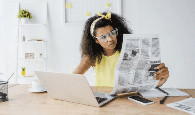 Woman wearing glasses reads a newspaper at a desk with a laptop, smartphone, and notebook. She appears focused.