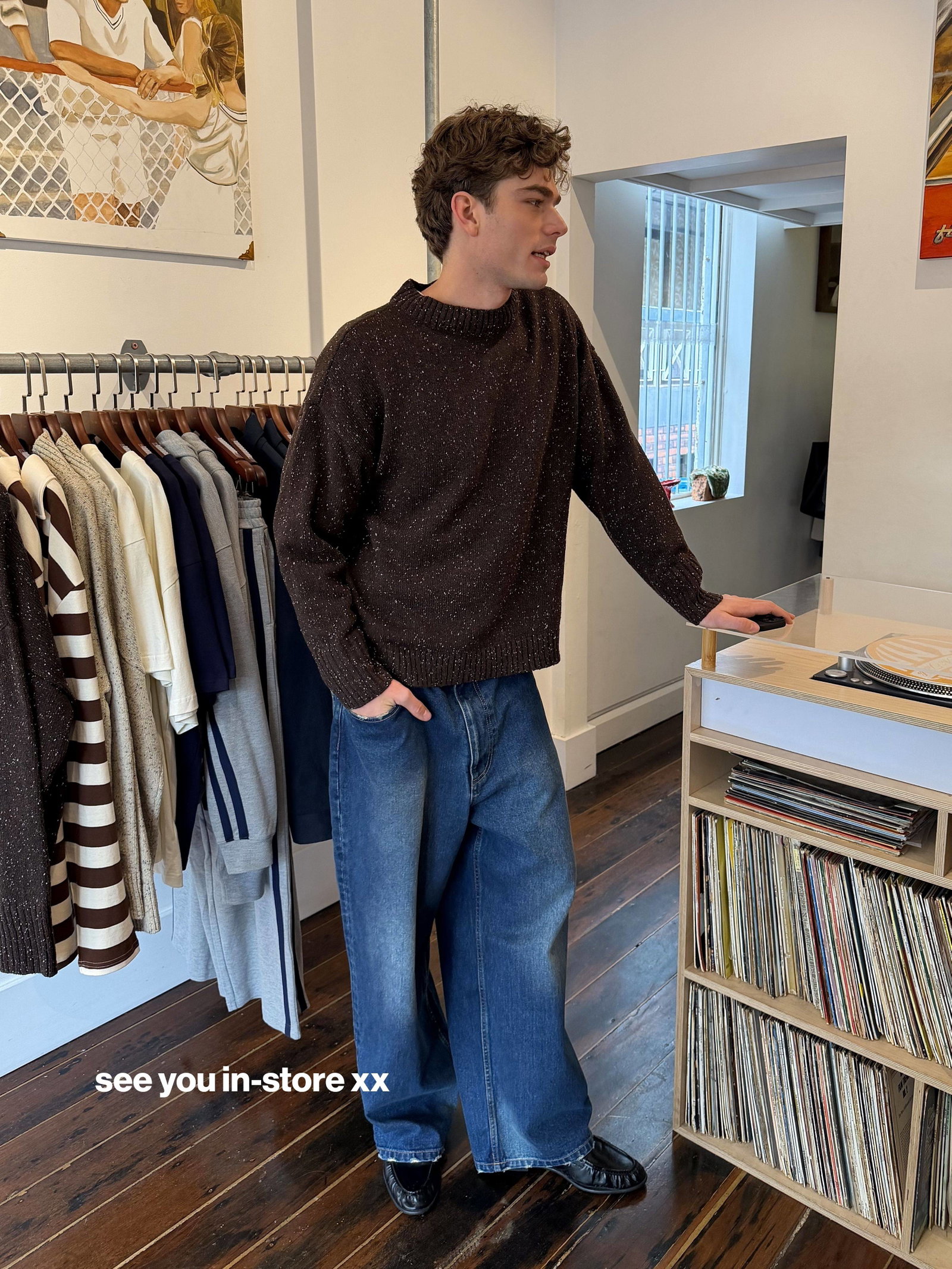 A young man with curly brown hair stands in a retail space, wearing a dark brown speckled sweater and wide-leg blue jeans, with his hand in his pocket...
