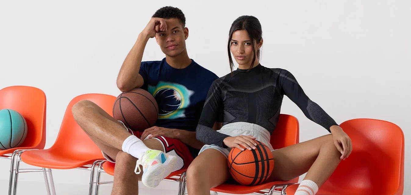 Zwei Personen sitzen auf orangen Plastikstühlen, jeweils einen Basketball haltend. Der eine trägt ein schwarzes enges Oberteil und Shorts, der andere ein buntes T-Shirt und Shorts.