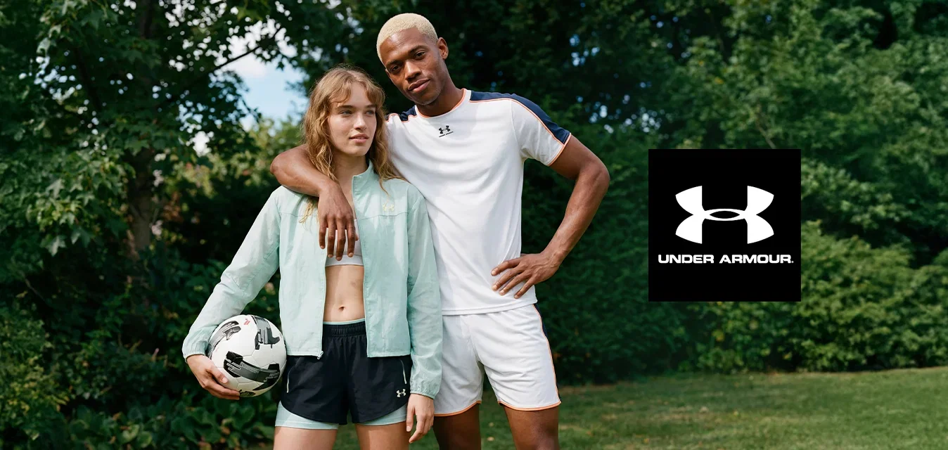 Zwei junge Erwachsene stehen auf Gras; eine Person hält einen Fussball, die andere hat einen Arm um sie gelegt. Beide tragen Sportkleidung, im Hintergrund sind Bäume.