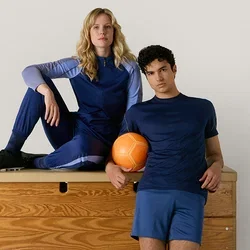 Zwei Personen in blauer Sportbekleidung posieren mit einem orangefarbenen Ball auf einem hölzernen Turnkasten vor einem schlichten hellen Hintergrund.