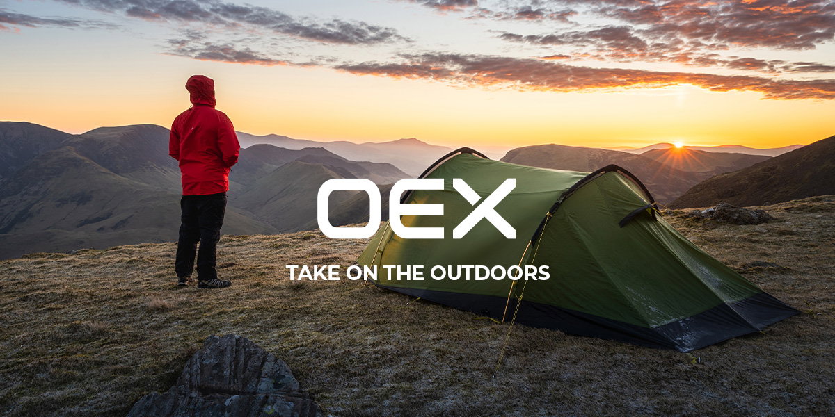 A wide outdoor scene at sunrise in a mountainous landscape. In the foreground on the right, a green camping tent is set up on a grassy, rocky hill. To the left, a person wearing a bright red hooded jacket and dark trousers stands facing away, looking out over layered hills and valleys. The sky is filled with soft clouds lit by orange and golden sunlight, with the sun just rising near the horizon on the right side of the image. The terrain appears rugged and open, evoking a remote hiking or camping location. Centered over the image is a white logo reading “OEX,” with the tagline “TAKE ON THE OUTDOORS” displayed below it.