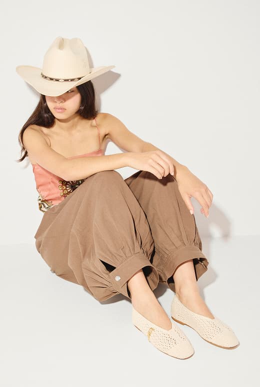 woman sitting in cowboy hat with pink silk top and brown pants wearing crochet ballet flats