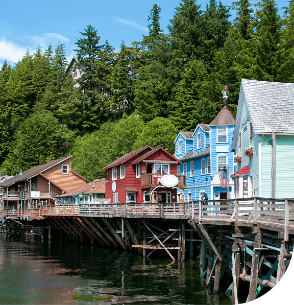 Colorful wooden buildings on stilts by a waterfront with lush trees in the background. Ketchikan, Alaska