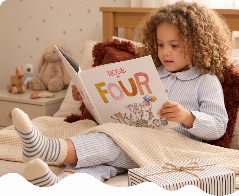 A young girl sitting in bed reading a personalised birthday book titled "Rose is Four" surrounded by soft toys and wrapped gifts