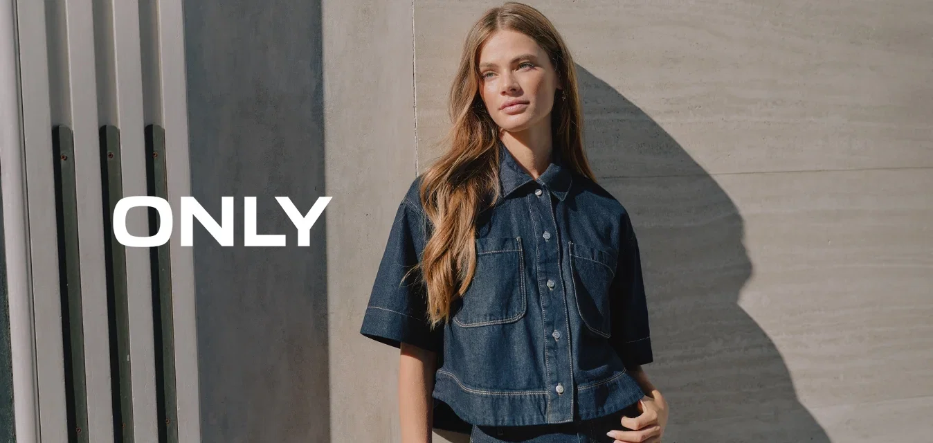 Junge Frau mit langem Haar, die vor einer Betonwand steht und ein dunkles Denim-Kurzarmhemd sowie passende hochtaillierte Jeans trägt.