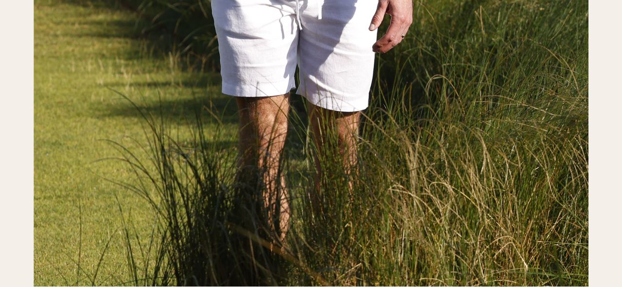 Close-up of legs in white linen shorts on grass. Westport Black Cole Linen-Blend Cabana Shirt label.
