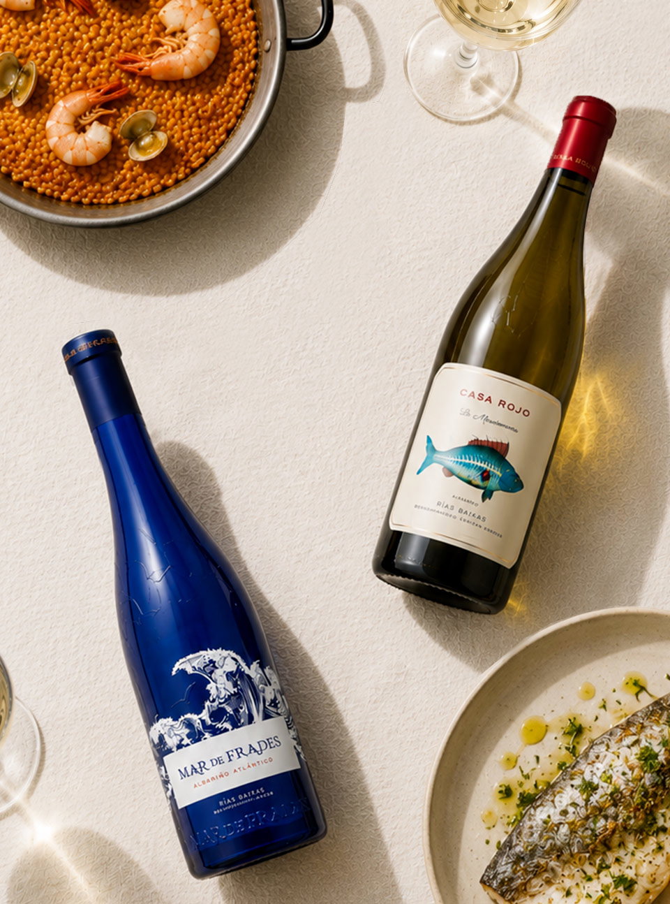 Top-down view of two white wine bottles and glasses beside prawn paella and grilled fish on a textured table