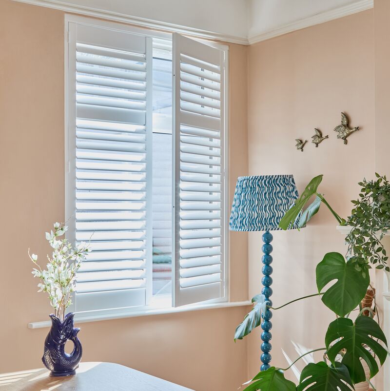 Full height white shutters installed on a window in a soft peach living room with potted plants