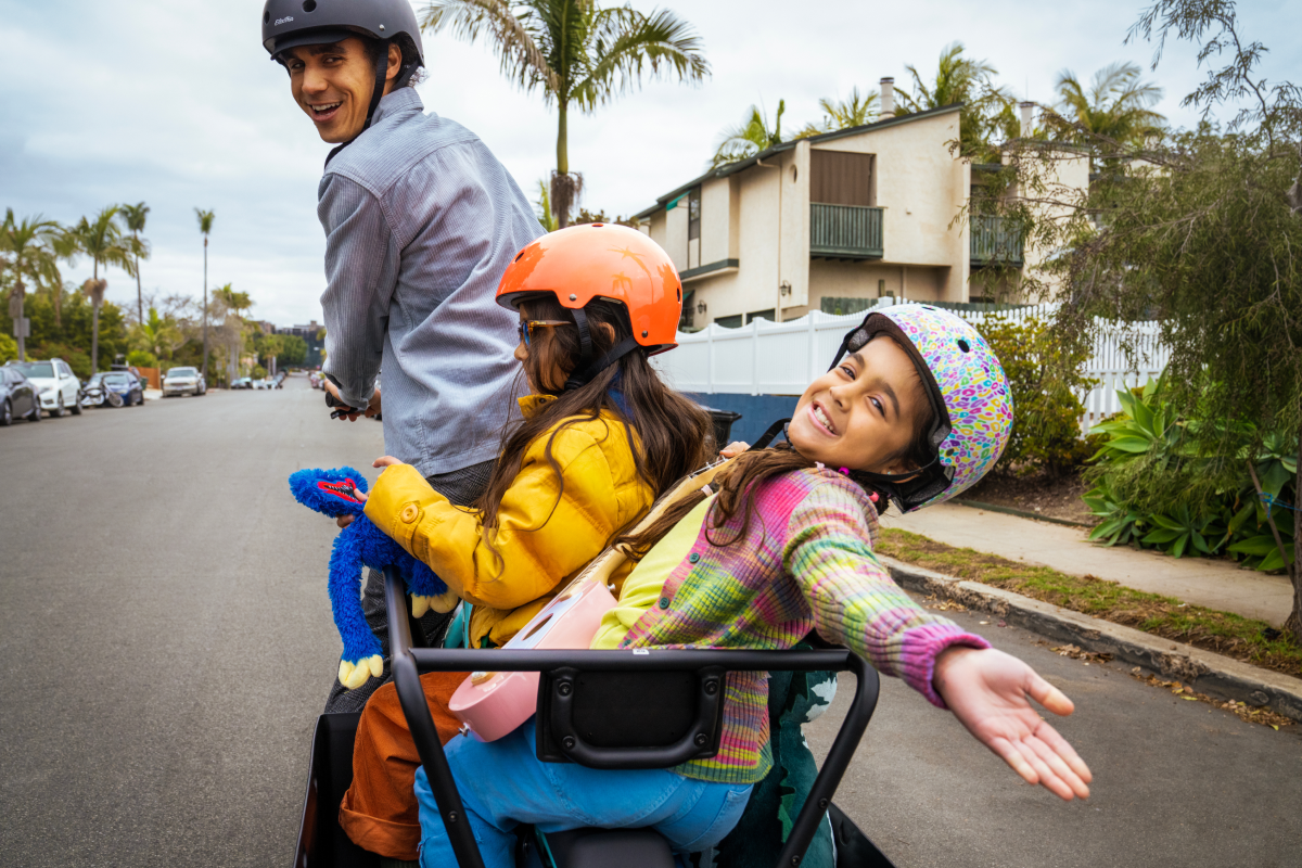 A man with a helmet drives a vehicle carrying two smiling girls in helmets, with palm trees and houses lining a suburban street in the background.