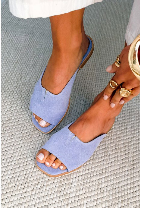 Woman's light-blue suede open-toe flat sandals on bare feet with white pedicure and gold rings, photographed on a textured woven rug