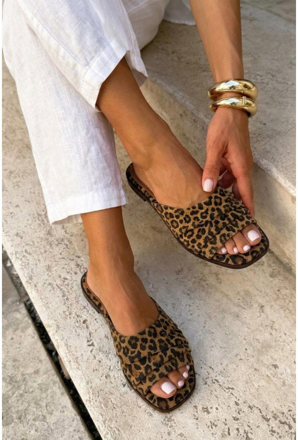 Woman wearing leopard-print slide sandals and white linen trousers, pale-pink pedicure visible, gold bangles, seated on stone steps