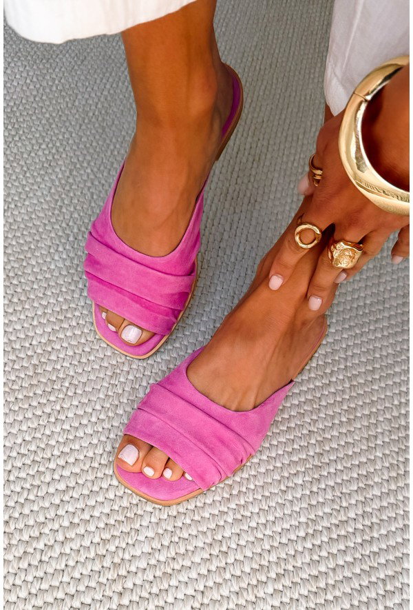 Woman's feet in pink suede open-toe flat sandals on a textured grey rug, white pedicure and gold rings