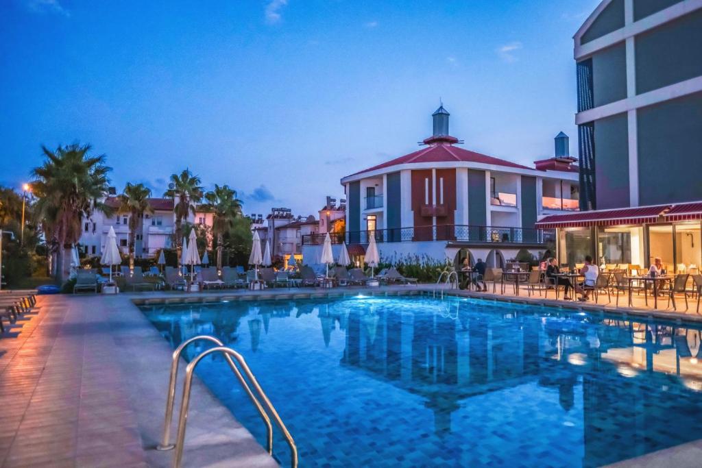 Hotel swimming pool at dusk with lit terrace, palm trees, sun loungers and guests dining beside reflective blue water